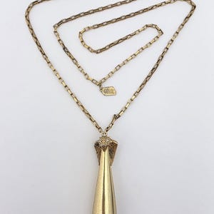 Op de afbeelding: Een goudkleurige ketting met een lange ketting en een druppelvormige hanger. De hanger heeft een cobra-kop ontwerp. De ketting is gemaakt van rechthoekige schakels en heeft een klein label met "GOVIL".