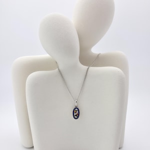 May include: A silver necklace with a dark blue oval pendant. The pendant features a floral design with white, red, and yellow accents. The necklace is displayed on a white mannequin.