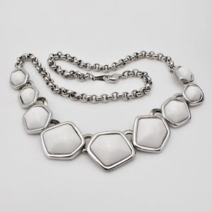 Puede incluir: Un collar plateado con colgantes blancos de forma geométrica. El collar tiene una cadena con cierre de mosquetón. Los colgantes están delineados en plata y tienen un acabado liso y brillante. El collar se muestra sobre un fondo blanco.