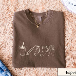 May include: Espresso-colored t-shirt with a white graphic of a drink, syringe, socks, and fries. A gold necklace and white sneakers are also visible. The shirt is folded on a fluffy, peach-colored rug.
