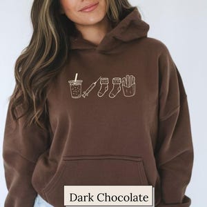 May include: Dark chocolate brown hoodie with a front pocket and a drawstring hood. The hoodie features a white line drawing of a drink, syringe, socks, and fries. The text "Dark Chocolate" is at the bottom.
