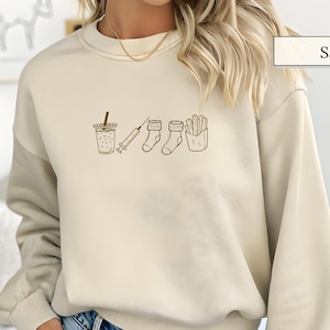May include: Sand-colored sweatshirt with a brown graphic of a drink, syringe, socks, and fries. The sweatshirt has long sleeves and a crew neck. The word "Sand" is in a rectangular box in the upper right corner.