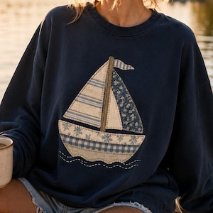 May include: A navy blue crewneck sweatshirt featuring a large, central applique of a sailboat. The sailboat is constructed from various fabric patches in shades of blue, cream, and white, showcasing striped, floral, and gingham patterns.