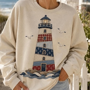 May include: A beige crewneck sweatshirt featuring a large, detailed graphic of a lighthouse. The lighthouse design incorporates various patterns including red and white gingham, blue with white anchors, and stripes. Three seagulls fly nearby.