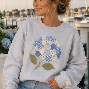 Puede incluir: Una sudadera gris claro de cuello redondo con una gran aplicación central de una flor de hortensia. La flor está compuesta por varios parches de tela azul, blanca y crema con patrones de cuadros, rayas y flores, junto con hojas verdes.