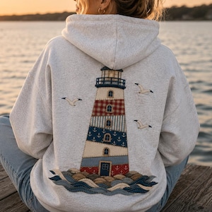 May include: A light grey heathered hoodie featuring a large embroidered lighthouse design on the back. The lighthouse is composed of various patterned sections including red gingham, blue anchors, and stripes, surrounded by three flying seagulls.