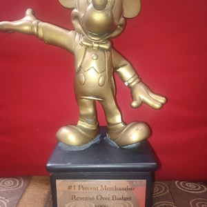 May include: A gold-colored Mickey Mouse statue on a black base. The statue is in a welcoming pose, with one arm extended. The base has a plaque that reads "#1 Percent Merchandise Revenue Over Budget 1998."