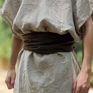 May include: A light grey linen tunic with short sleeves and a dark brown belt. The tunic is loose-fitting and appears to be made of a soft, breathable fabric. The belt is wrapped around the waist, creating a defined silhouette. The overall style is simple and rustic.