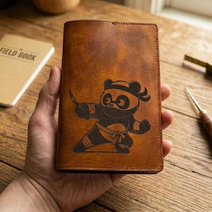 May include: A brown leather wallet with a ninja panda design. The wallet is held in a hand and features a detailed illustration of a panda in a ninja outfit. The wallet is rectangular and has a stitched border.