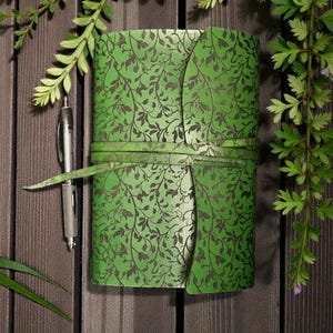 May include: A green journal with a leaf pattern and a matching tie closure. A silver pen rests beside the journal on a wooden surface. The journal's cover is a vibrant green with a darker leaf design.