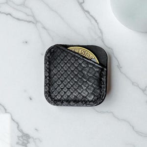 May include: A square, black leather coin holder with a textured, snake-skin pattern. A gold-colored coin with the word "MENTO" is partially visible in the pocket. The holder is on a white marble surface.