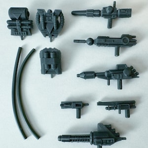 Könnte beinhalten: Eine Sammlung grauer Spielzeugwaffen und Zubehörteile aus Kunststoff, darunter verschiedene Blaster, Rucksäcke und Schläuche. Die Teile sind mit mechanischen Designs detailliert, was auf ein Science-Fiction- oder Action-Thema hindeutet. Die Artikel sind auf einer weißen Oberfläche angeordnet.