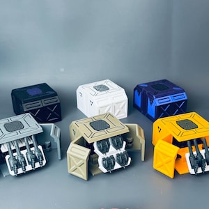 Könnte beinhalten: Eine Sammlung farbenfroher, miniaturisierter, futuristisch aussehender Boxen. Die Boxen sind in verschiedenen Farben erhältlich, darunter Schwarz, Weiß, Blau, Beige und Gelb. Einige Boxen haben offene Fächer mit detaillierten mechanischen Komponenten.