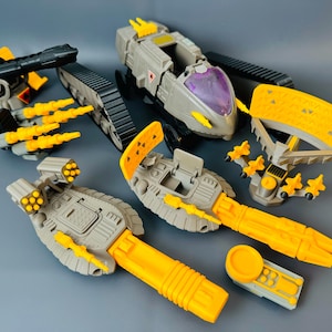 Könnte beinhalten: Eine Sammlung grauer und gelber Spielzeugfahrzeuge, darunter Panzer, Raketenwerfer und eine Radarschüssel. Die Spielzeuge haben ein futuristisches Design mit verschiedenen Details, wie z. B. Raketenkapseln und einem lila Cockpit. Die Spielzeuge sind aus Kunststoff.