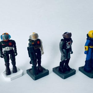 Może przedstawiać: Cztery figurki akcji w stylu vintage przedstawiające futurystycznych żołnierzy. Każda figurka stoi na małej podstawie i nosi hełm. Figurki są w różnych kolorach, w tym czarnym, niebieskim, szarym i żółtym. Figurki stoją prosto.