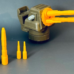 Könnte beinhalten: Eine olivgrüne Militärkanzel mit zwei orangefarbenen Läufen und mehreren orangefarbenen Projektilen. Der Turm hat einen hellblauen Knopf. Die Projektile sind in verschiedenen Größen gehalten, alle in leuchtendem Orange.