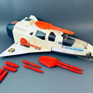 Könnte beinhalten: Ein Vintage-Starcom-Raumschiff-Spielzeug in Creme, Orange und Marineblau. Das Spielzeug hat ein Cockpit, Flügel und mehrere rote Raketenzubehörteile. Der Text "STARCOM" ist auf der Seite des Schiffs zu sehen.
