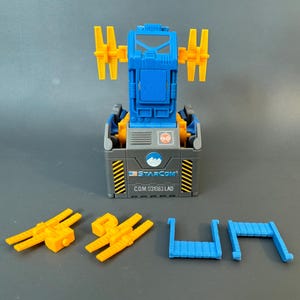 Könnte beinhalten: Ein blau-gelbes Starcom-Spielzeugset. Die Hauptkomponente ist eine graue Box mit blauem Deckel und gelben Akzenten. Zusätzliche gelbe und blaue Teile sind enthalten. Auf der Box steht "STARCOM" und "C.O.M. 031083 LAD".