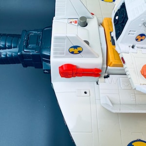 Könnte beinhalten: Ein weiß-gelbes Spielzeugraumschiff mit einer dunkelblauen Kanone und roten und orangefarbenen Akzenten. Das Spielzeug hat ein detailliertes Design mit verschiedenen Paneelen, Aufklebern und einem Cockpitbereich. Das Spielzeug ist aus Kunststoff.