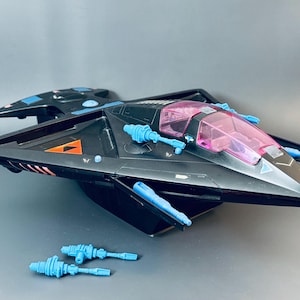 Könnte beinhalten: Schwarzes Spielzeugraumschiff mit pinkfarbenem Cockpit und blauen Akzenten. Das Spielzeug hat Flügel und mehrere blaue Blaster. Ein orangefarbenes Dreieck ist an der Seite sichtbar. Zwei zusätzliche blaue Blaster befinden sich vor dem Schiff.