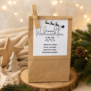 Könnte beinhalten: Eine braune Papiertüte mit einem weißen Etikett, das eine schwarze Grafik von einem Schlitten mit Rentieren sowie den Text 15 Minuten Weihnachten in der Tüte, Cappuccino kochen, Kerze anzünden, Geschichte lesen, Schokolade genießen zeigt, befestigt mit einer Holzklammer.