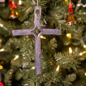 May include: A rustic, gray-toned cross Christmas ornament hanging on a decorated tree. The cross is made of a textured material and has a hole at the top for hanging. The background features a green Christmas tree with lights and other ornaments.