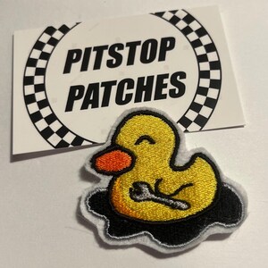 May include: Yellow embroidered iron-on patch of a cartoon duck with a wrench in its hand, sitting in a puddle of oil. The patch is labelled "Pitstop Patches".