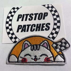 May include: Embroidered patch of a smiling white cat with pink ears and red details, holding a chequered flag. The cat is set against a yellow background. A sticker above the patch reads "PITSTOP PATCHES" in black.