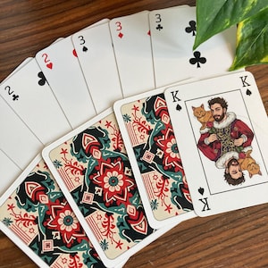 K&ouml;nnte beinhalten: Ein Kartenspiel mit einem einzigartigen Design. Die Karten zeigen einen K&ouml;nig mit einem Mann, der zwei Katzen h&auml;lt, und die R&uuml;ckseiten der Karten haben ein rotes, schwarzes und blaugr&uuml;nes Blumenmuster. Die Farben sind auf den Bildkarten sichtbar.