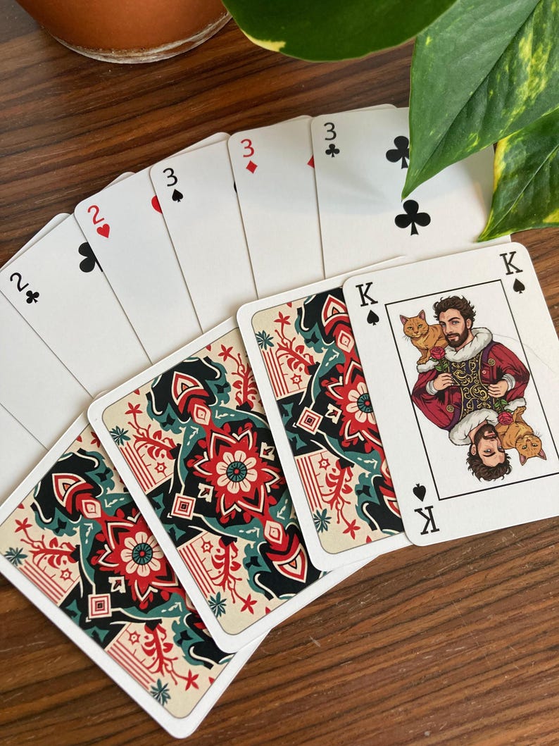K&ouml;nnte beinhalten: Ein Kartenspiel mit einem einzigartigen Design. Die Karten zeigen einen K&ouml;nig mit einem Mann, der zwei Katzen h&auml;lt, und die R&uuml;ckseiten der Karten haben ein rotes, schwarzes und blaugr&uuml;nes Blumenmuster. Die Farben sind auf den Bildkarten sichtbar.