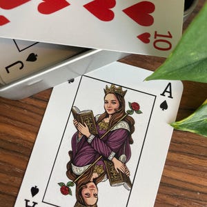 K&ouml;nnte beinhalten: Ein dekoratives Kartenspiel. Das Pik-Ass zeigt eine Frau in einem lila Kleid, die ein Buch und eine Rose h&auml;lt. Das Spiel enth&auml;lt die 10 der Herzen und den Buben Pik. Die Karten liegen auf einer Holzoberfl&auml;che.