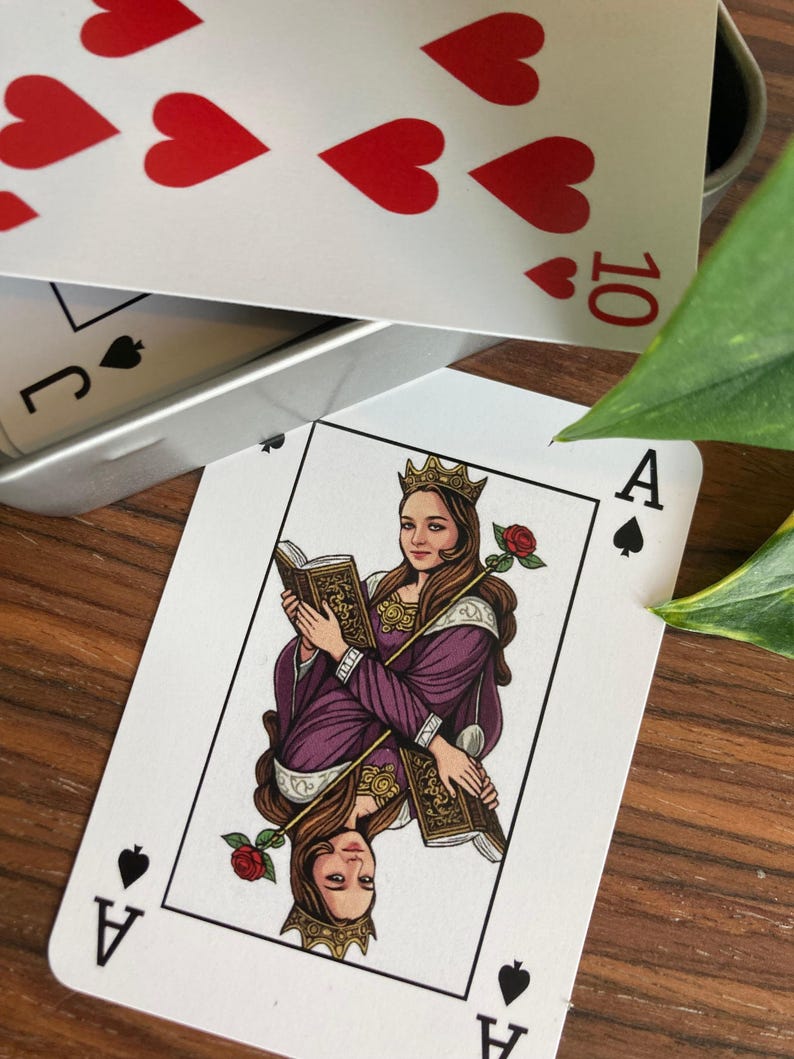 K&ouml;nnte beinhalten: Ein dekoratives Kartenspiel. Das Pik-Ass zeigt eine Frau in einem lila Kleid, die ein Buch und eine Rose h&auml;lt. Das Spiel enth&auml;lt die 10 der Herzen und den Buben Pik. Die Karten liegen auf einer Holzoberfl&auml;che.