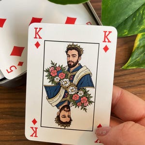 K&ouml;nnte beinhalten: Eine Spielkarte mit dem K&ouml;nig der Karo. Die Karte zeigt einen gekr&ouml;nten Mann mit Blumen, in blauen und goldenen Gew&auml;ndern. Die Karte wird in einer Hand gehalten, mit einem Teilblick auf andere Karten im Hintergrund.