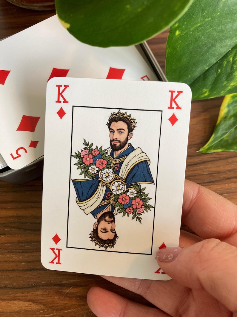 K&ouml;nnte beinhalten: Eine Spielkarte mit dem K&ouml;nig der Karo. Die Karte zeigt einen gekr&ouml;nten Mann mit Blumen, in blauen und goldenen Gew&auml;ndern. Die Karte wird in einer Hand gehalten, mit einem Teilblick auf andere Karten im Hintergrund.