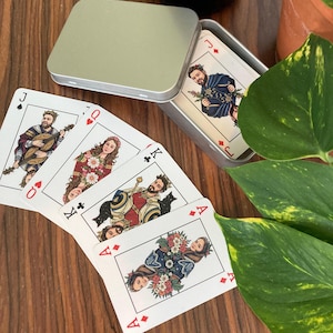 May include: A deck of playing cards with illustrated figures, including a jack, queen, king, and ace of diamonds. The cards are in a silver tin, with a bottle of Prosecco in the background. The cards are on a wooden surface.
