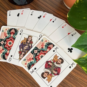 K&ouml;nnte beinhalten: Ein Kartenspiel auf einer Holzoberfl&auml;che aufgef&auml;chert. Die Karten zeigen individuelle Illustrationen einer K&ouml;nigin mit einer Katze und eines K&ouml;nigs mit zwei Katzen. Die Farben sind sichtbar, darunter Herz, Pik, Kreuz und Karo.