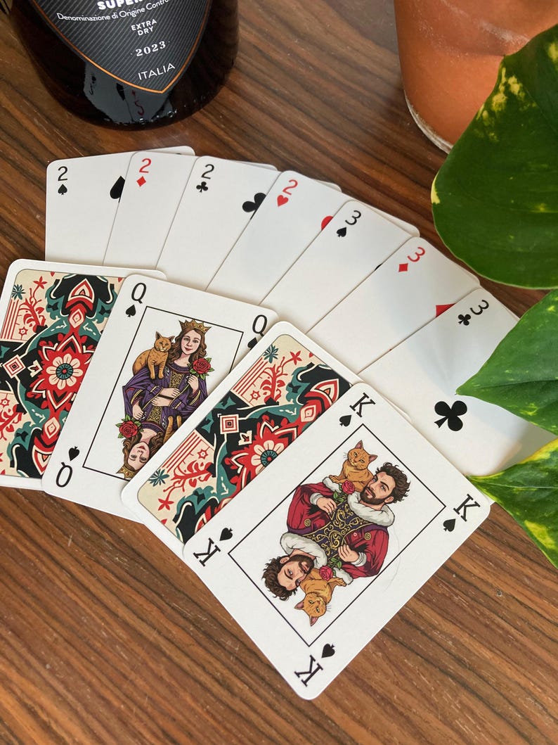 K&ouml;nnte beinhalten: Ein Kartenspiel auf einer Holzoberfl&auml;che aufgef&auml;chert. Die Karten zeigen individuelle Illustrationen einer K&ouml;nigin mit einer Katze und eines K&ouml;nigs mit zwei Katzen. Die Farben sind sichtbar, darunter Herz, Pik, Kreuz und Karo.