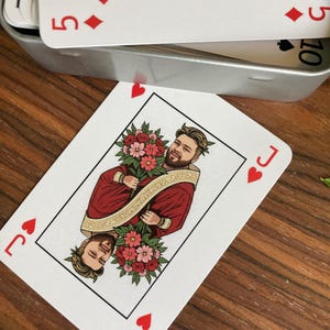 K&ouml;nnte beinhalten: Spielkarte, der Bube der Herzen, mit einem Portr&auml;t einer Person in roter Robe, die Blumen h&auml;lt. Die Karte hat rote Herzsymbole. Weitere Karten sind in einer Metallbox sichtbar.