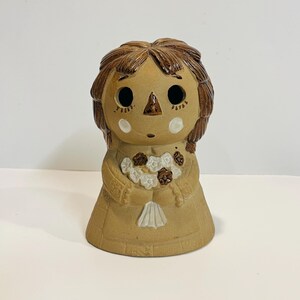 May include: A ceramic doll figurine with brown hair, a brown dress, and a white flower bouquet. The doll has a simple, painted face with brown eyes and a brown nose.