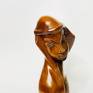 May include: A wooden sculpture of a woman's bust. The sculpture is carved from a dark brown wood and features a smooth, polished finish. The woman is depicted with a serene expression, wearing a hat. The sculpture is a decorative art piece.