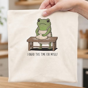 Könnte beinhalten: Eine cremefarbene Tragetasche mit einer grafischen Illustration eines grünen Frosches in einer Schürze, der an einem Holztisch steht und Teig knetet. Unter der Illustration steht der Text "I KNEAD THIS TIME FOR MYSELF" in schwarzer Schrift.