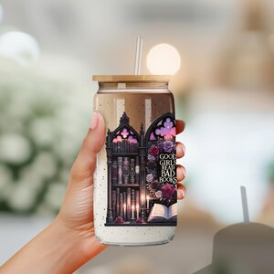 May include: A clear glass tumbler with a black and white graphic of a bookshelf filled with books. The graphic also includes a gothic archway, candles, and flowers. The tumbler has a bamboo lid and a straw. The text on the tumbler reads "Good girls read bad books."