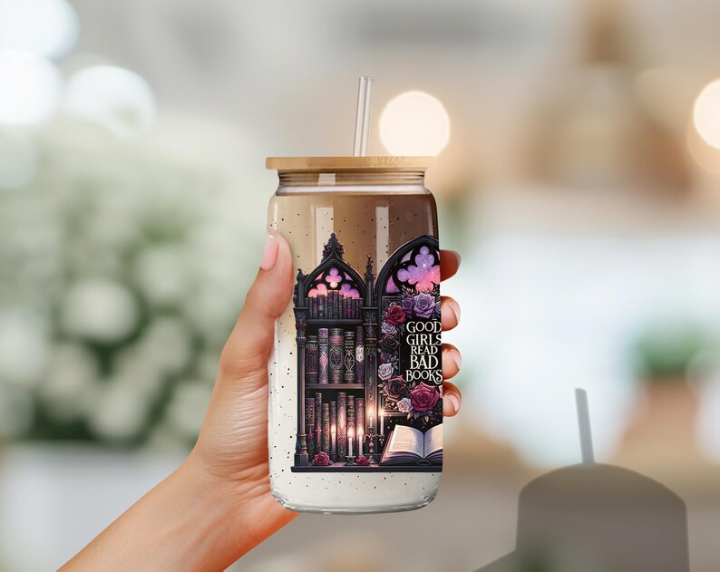 May include: A clear glass tumbler with a black and white graphic of a bookshelf filled with books. The graphic also includes a gothic archway, candles, and flowers. The tumbler has a bamboo lid and a straw. The text on the tumbler reads "Good girls read bad books."