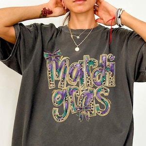 Op de afbeelding: Donkergrijs t-shirt met een kleurrijke "Mardi Gras"-afbeelding. Het ontwerp heeft paarse, blauwgroene en gouden accenten, inclusief strikdetails en een luipaardprint. Een casual, oversized t-shirt.