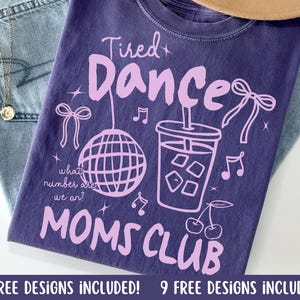 Puede incluir: Una camiseta morada con las palabras "Tired Dance Moms Club" en una fuente morada clara. La camiseta presenta ilustraciones de una bola de discoteca, una bebida con cubitos de hielo, notas musicales y cerezas. La camiseta está doblada sobre unos vaqueros azules.