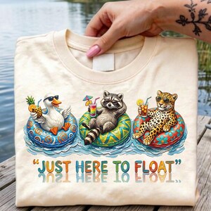 Könnte beinhalten: Cremefarbenes T-Shirt mit einem skurrilen Design einer Ente, eines Waschbären und eines Leoparden, die sich auf aufblasbaren Ringen im Wasser entspannen, jeder mit einem tropischen Getränk. Der Text "JUST HERE TO FLOAT" ist darunter gedruckt.