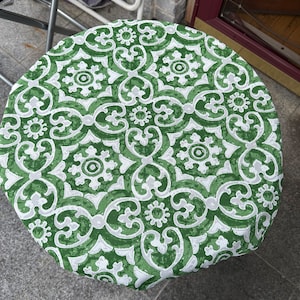May include: A round table cover with a green and white floral pattern. The design features a repeating motif of stylized flowers and swirling shapes. The cover is made of a fabric material and is placed on a round table.