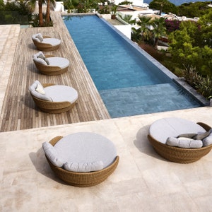 Peut inclure: Scène extérieure avec une longue piscine rectangulaire remplie d'eau bleue. Quatre chaises longues rondes en osier avec coussins et oreillers gris sont disposées sur une terrasse en bois et un patio en pierre. Des palmiers et de la verdure sont visibles.