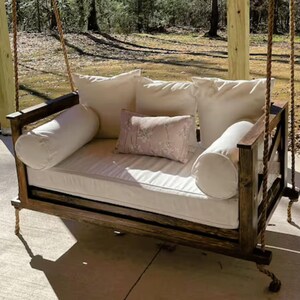 May include: A wooden swing bed with a white mattress and pillows. The swing is hanging from a porch ceiling and is made of dark wood.