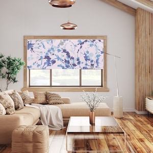 May include: A modern living room with a beige sectional sofa, wooden floors, and a large window with a patterned roller shade. The shade features a blue and white watercolor design. Copper pendant lights hang from the ceiling.